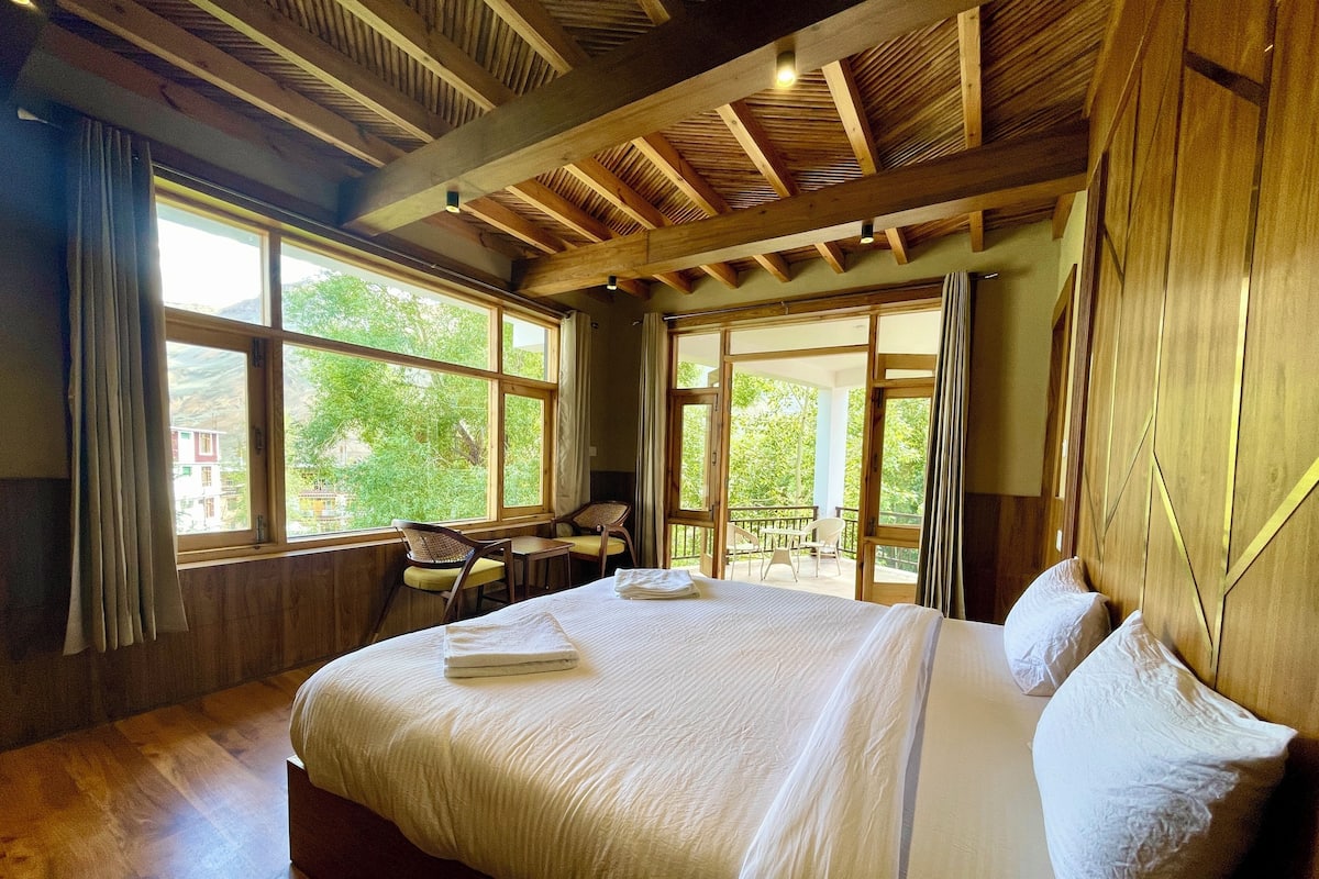 A bedroom with a large bed, wooden ceiling beams, and large windows with curtains.