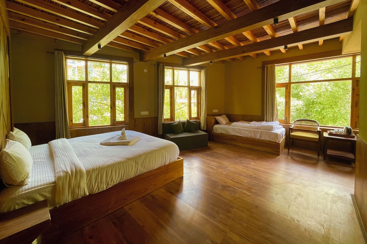 A room with two beds, a sofa, and a chair, with large windows offering a view of greenery.