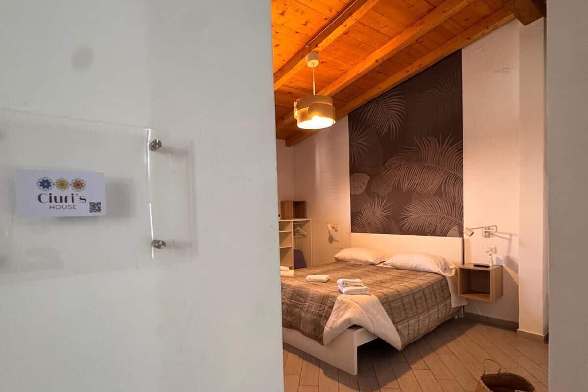 A bedroom with a wooden ceiling, a bed with a patterned bedspread, a bedside table with a lamp, and a wall with a leaf pattern.