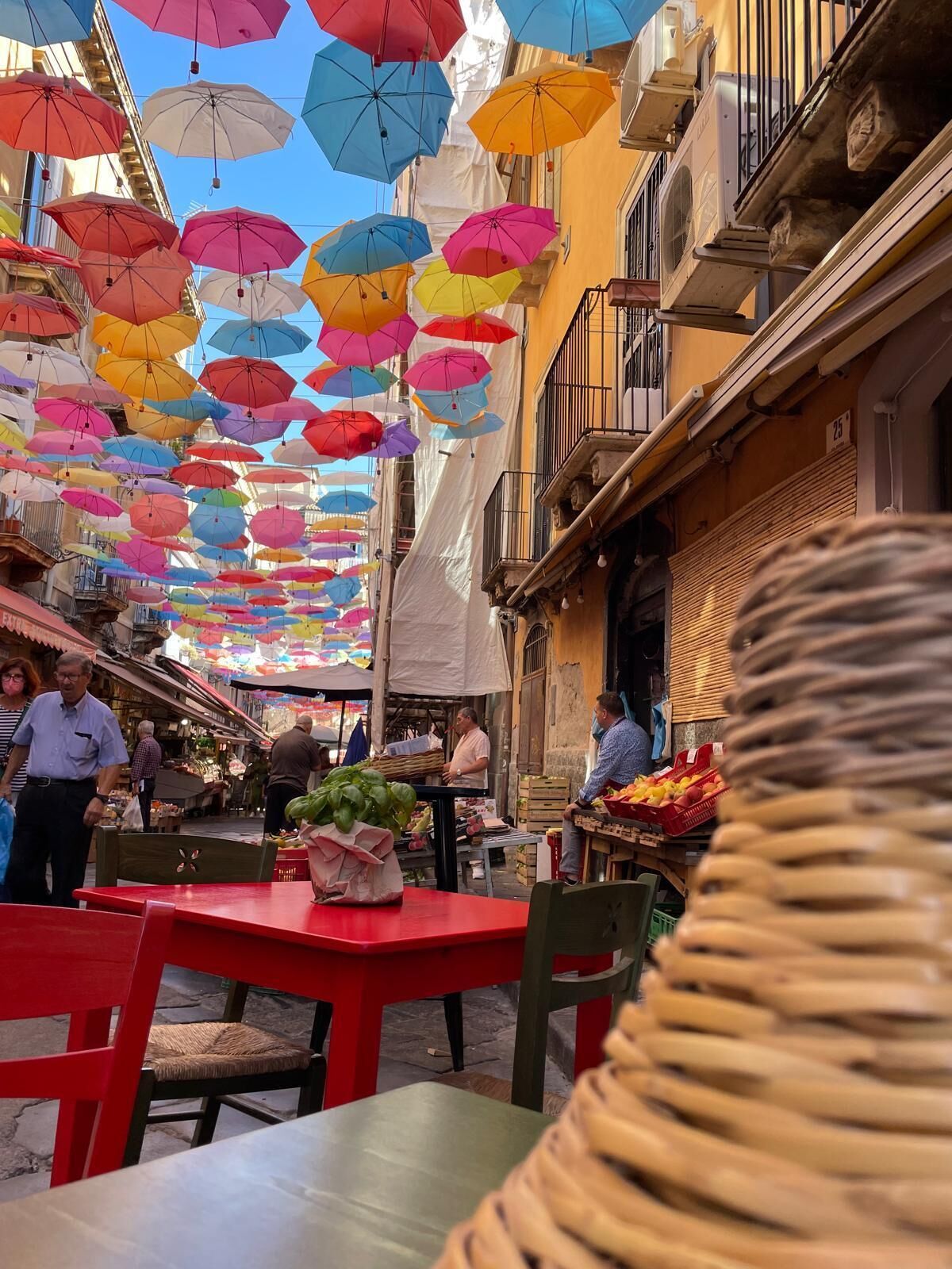 Une rue bordée de parasols colorés, un étal de marché proposant des produits frais et un immeuble doté d’un balcon.