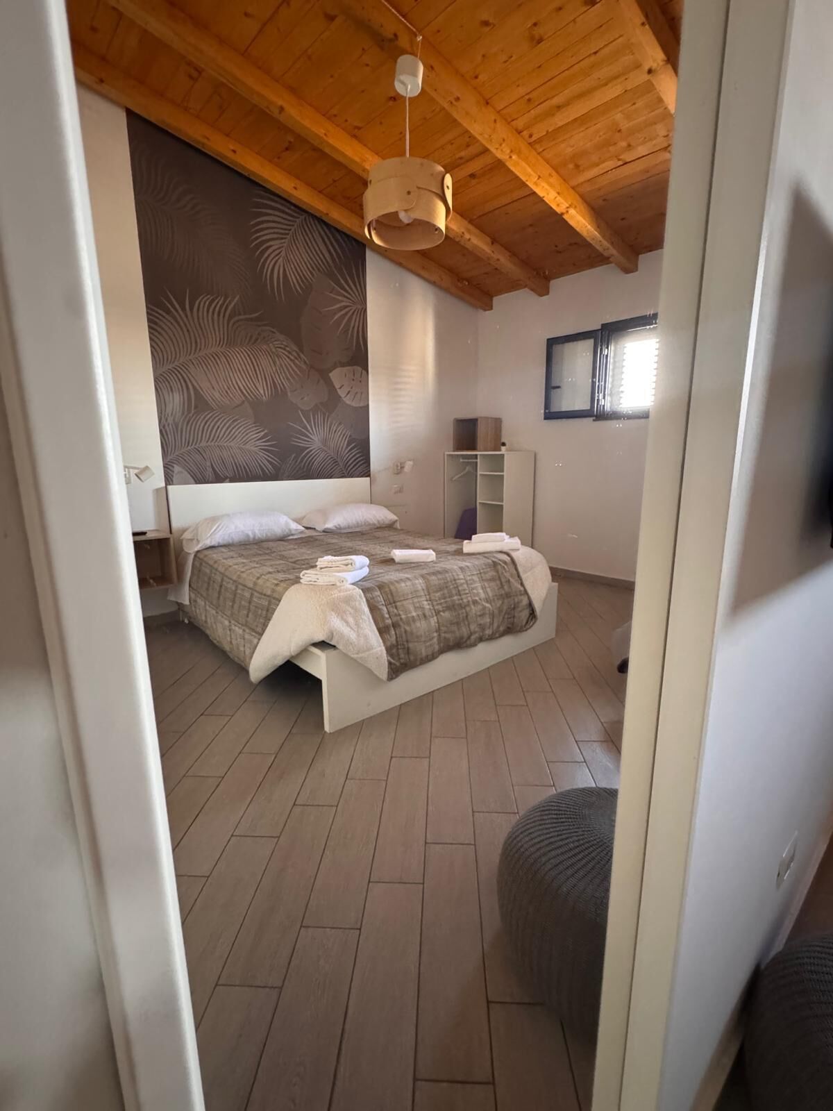 A bedroom with a wooden ceiling, a bed with a headboard, a wardrobe, and a window.