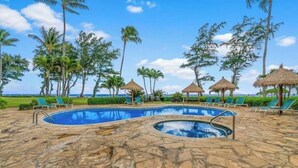 Zona de piscina con sillones de descanso y sombrillas de paja, rodeada de palmeras y un cielo despejado.