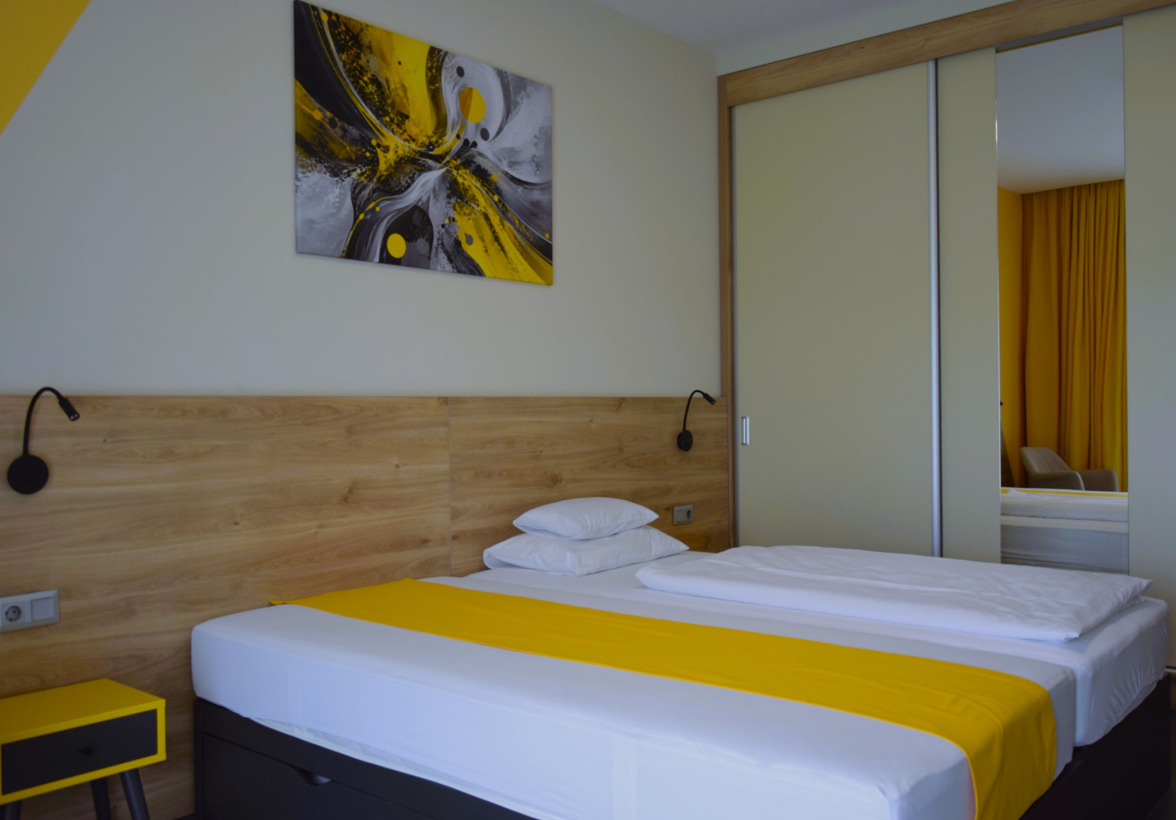 A hotel room with a wooden headboard, a bed with white bedding and a yellow accent, a bedside table, and a wall-mounted painting.