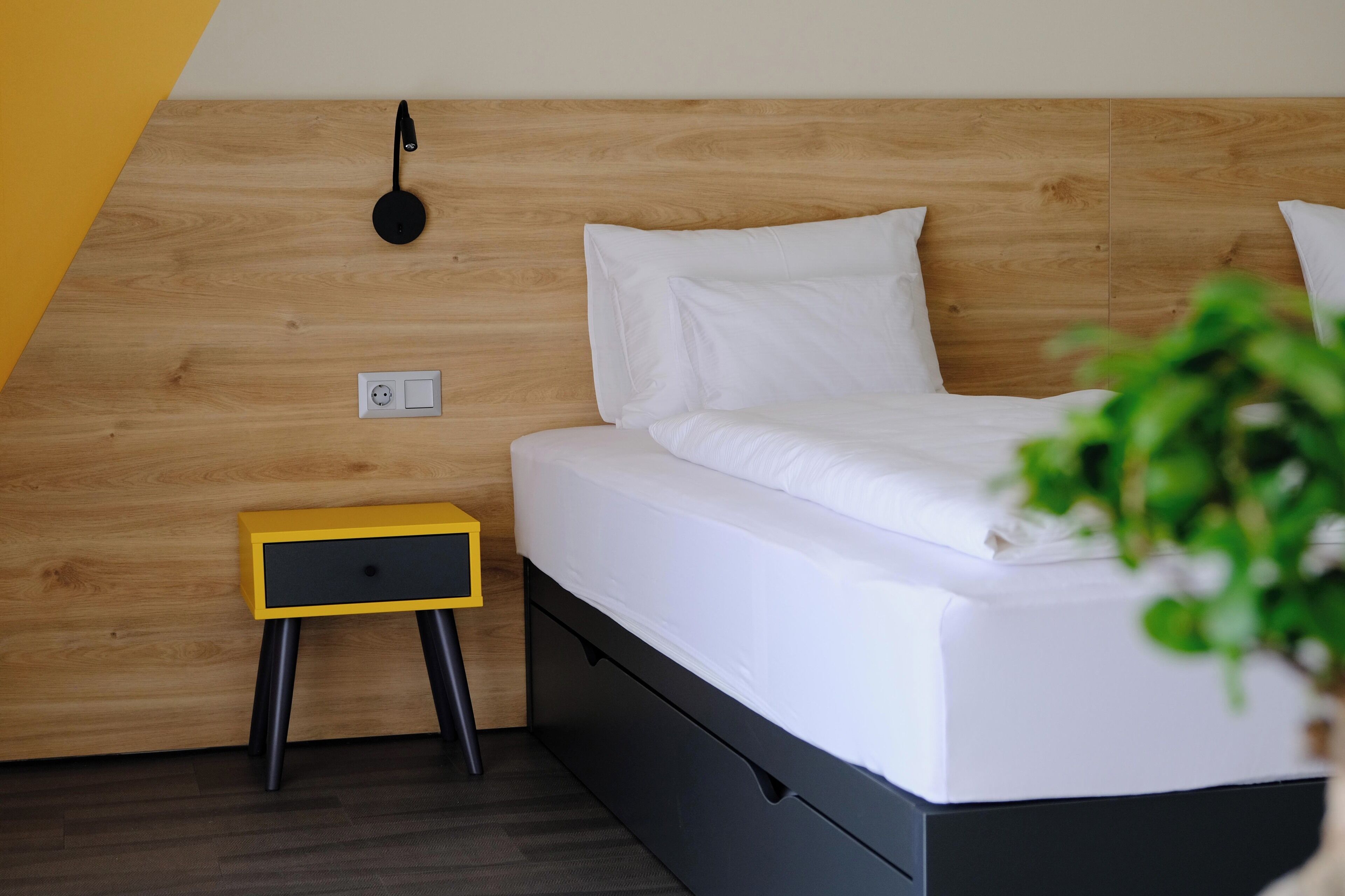 A neatly made bed with white linens, a small bedside table with a drawer, and a wall-mounted lamp.