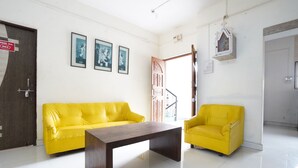 Una habitación luminosa con un sofá amarillo, una mesa de centro de madera y un sillón amarillo pequeño. Hay cuadros enmarcados en la pared y una puerta con un letrero.