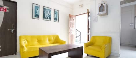 A bright room with a yellow sofa, a wooden coffee table, and a small yellow armchair. There are framed pictures on the wall and a door with a sign.