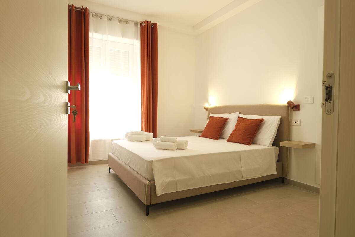 A neatly made bed with white linens and two pillows, a bedside table with a lamp, and a window with curtains.