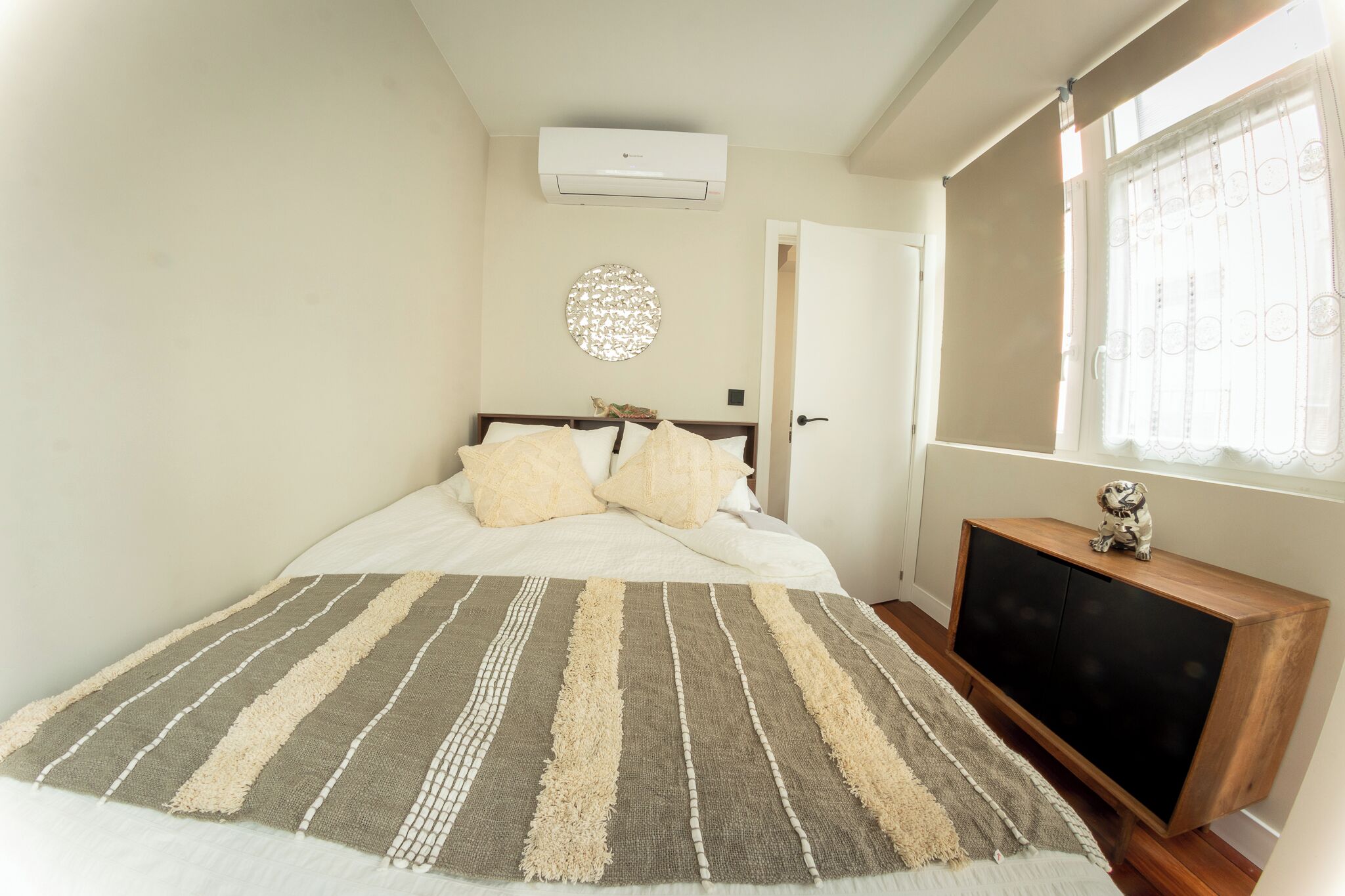 Une chambre à coucher comprenant un lit, une table de chevet, un climatiseur fixé au mur, un objet décoratif rond et une fenêtre avec un rideau.