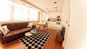 Een moderne woonkamer met een bruine bank, een ronde salontafel en een zwart-wit vloerkleed. Er is een keuken met witte kasten en een magnetron.