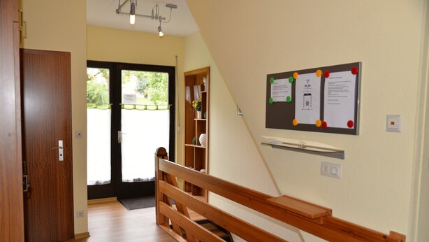 Un couloir avec un banc en bois, un tableau d’affichage où sont accrochées des annonces, et une porte qui donne sur une pièce.