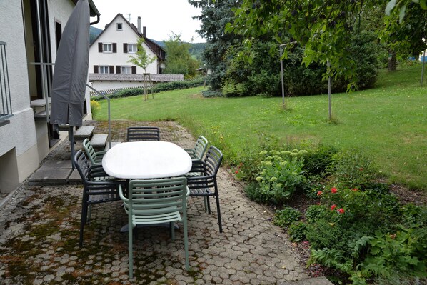 Eine Terrasse mit Tisch und Stühlen, einem schattenspendenden Sonnenschirm und einem Garten mit verschiedenen Pflanzen und Blumen.