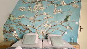 A bedroom with a bed, two pillows, a blue wall with a floral pattern, and a nightstand with a lamp. - Villa Medena (Mayenne)