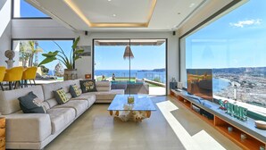A modern living room with a large sofa, a coffee table, and a TV on a wooden shelf. There is a large window with a view of the city and the sea. - Villa Bali by Marhen Homes (Javea / Xàbia)