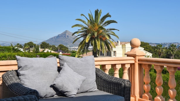 Ein Korbsessel mit grauen Kissen auf einem Balkon im Freien mit Blick auf einen Berg und Palmen.