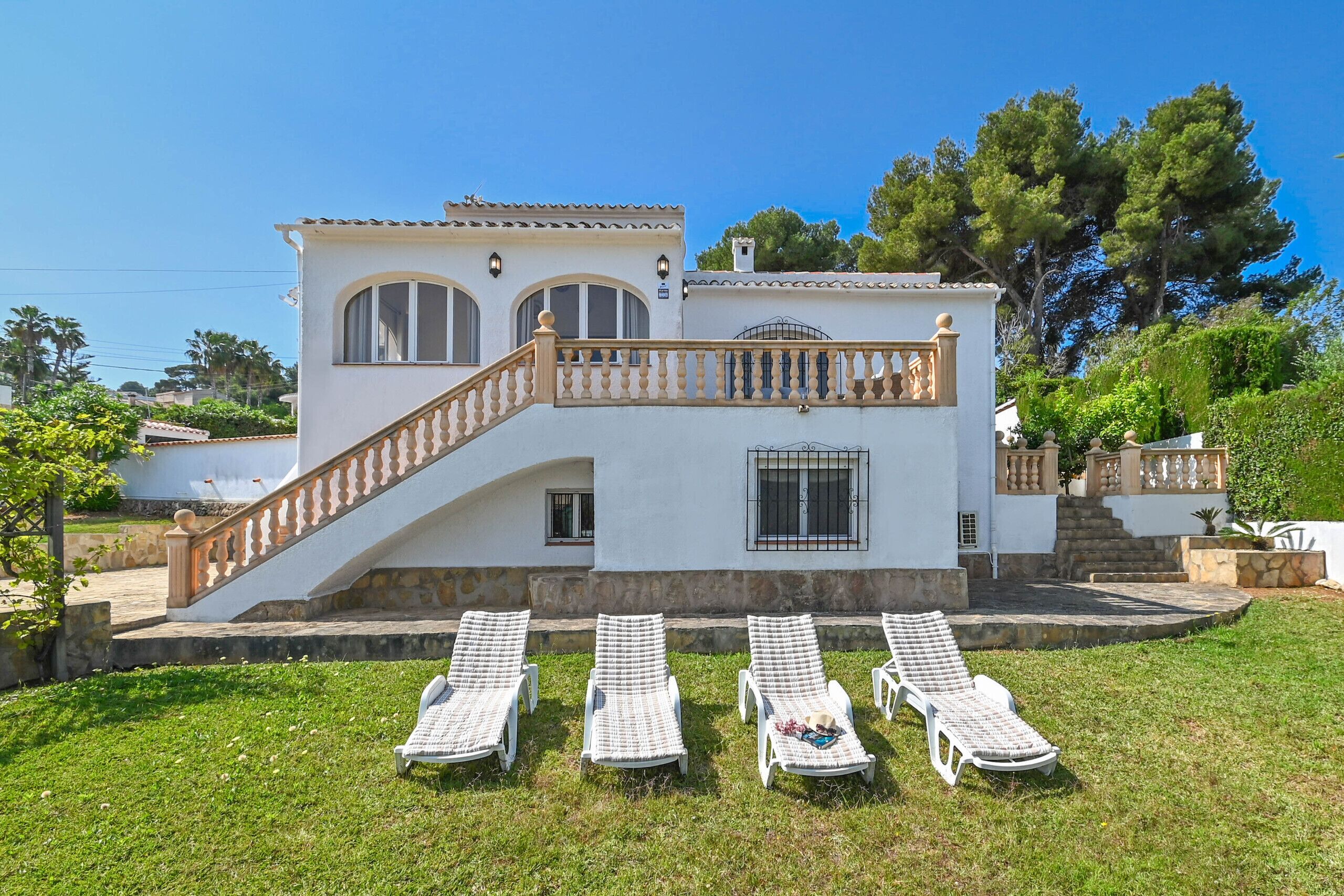 A two-story house with a white exterior, a wooden staircase, and a balcony. There are four lounge chairs in the front yard.