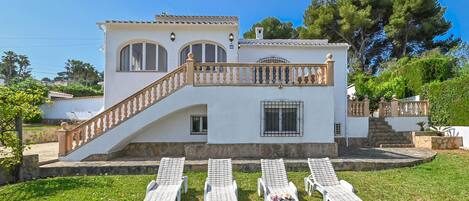 A two-story house with a white exterior, a wooden staircase, and a balcony. There are four lounge chairs in the front yard.