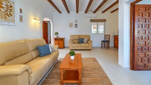 Ein Wohnzimmer mit beiger Couch, Couchtisch aus Holz und einem beigen Sessel. Es gibt einen Holzschrank und einen kleinen Tisch mit einer Pflanze. Der Raum hat Holzbalken an der Decke und eine Tür mit Muster.