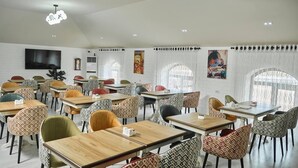A dining area with multiple tables and chairs, a flat-screen TV mounted on the wall, and a large window with curtains. - Siab Boutique Hotel (Samarkand)