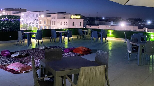 An outdoor seating area with tables and chairs under a large umbrella, overlooking a building with a sign that reads "oasis." - Siab Boutique Hotel (Samarkand)