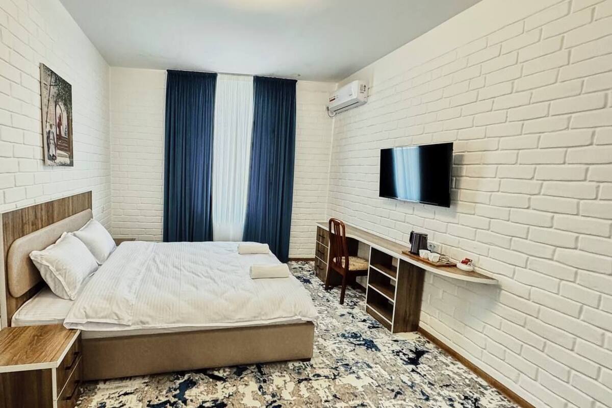 A modern hotel room with a brick-patterned wall, a bed with a wooden headboard, a desk with a chair, a flat-screen TV, and a large window with blue curtains.