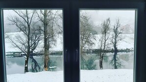 Une vue à travers une fenêtre sur un paysage enneigé, avec des arbres et une étendue d’eau.