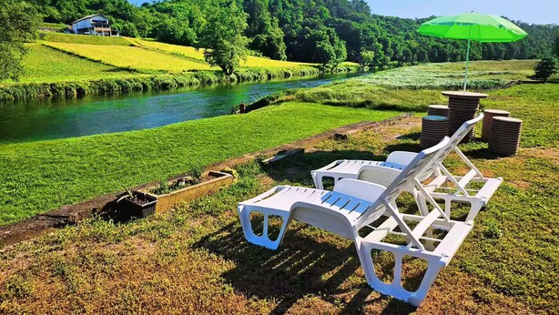 Un cadre au bord de la rivière, avec une chaise et un parasol, une petite barque et un bâtiment en arrière-plan.