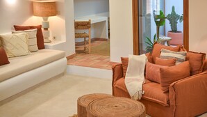 A living room with a white sofa, an orange armchair, and a woven coffee table. There is a lamp on a side table, and a potted plant is visible through the window. - Casa Paraiso by Marhen Homes (Javea / Xàbia)