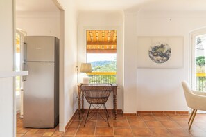 A kitchen with a refrigerator, a dining table with a chair, a window with a view, and a wall-mounted lamp.