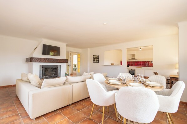 A modern living room with a fireplace, a dining area with white chairs and a table set for four, and a kitchen visible through an archway.