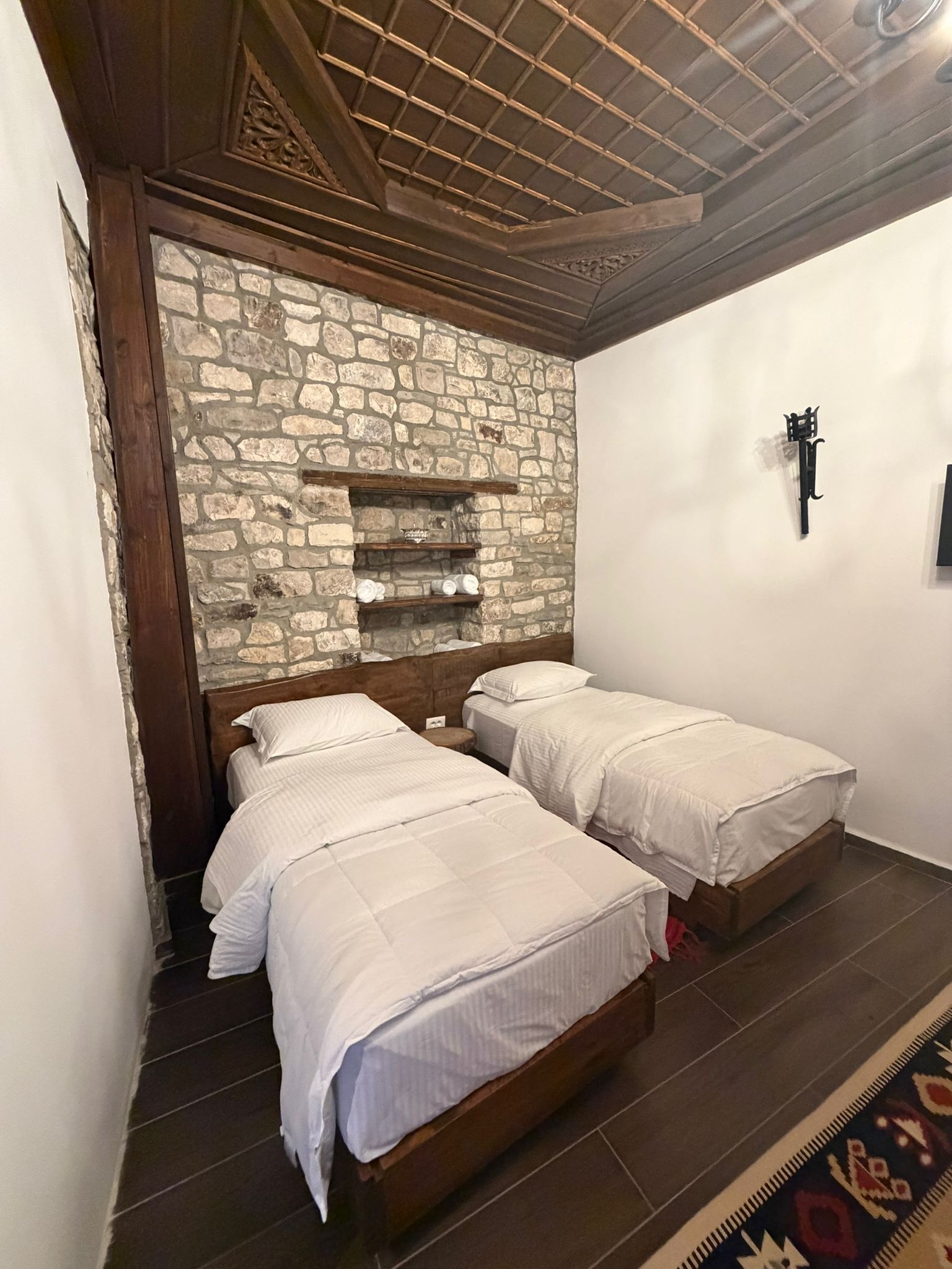 A bedroom with two beds, a stone wall, wooden beams, and a patterned rug.
