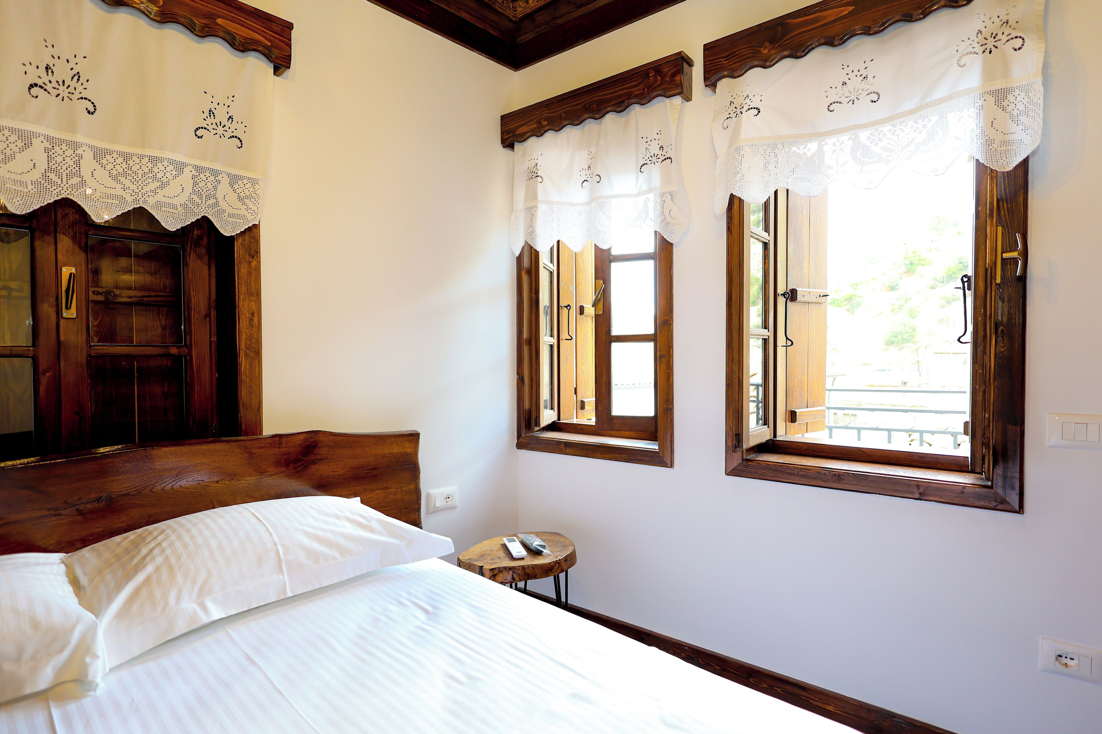 Une chambre à coucher avec une tête de lit en bois, un lit recouvert de draps blancs, une petite table ronde avec une télécommande, et trois fenêtres avec des cadres en bois et des rideaux blancs en dentelle.