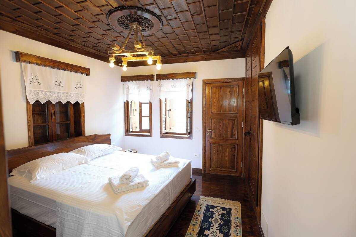 Un dormitorio con cama de madera, ropa de cama blanca, una araña y un televisor montado en la pared.
