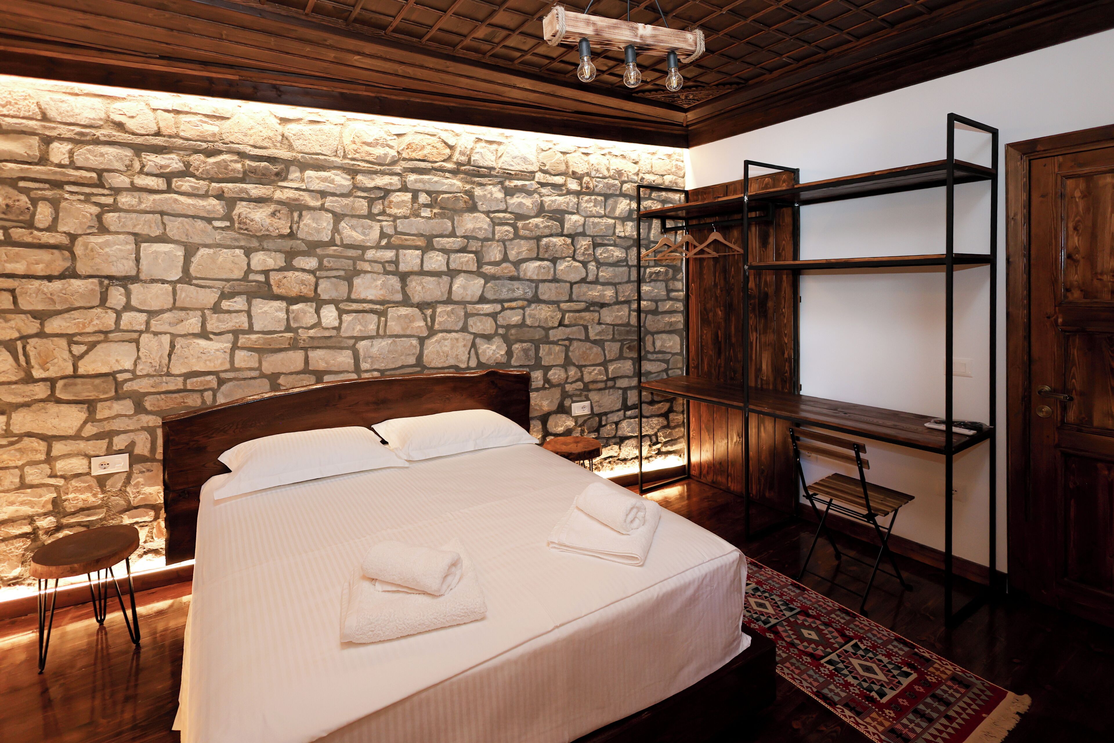 A bedroom with a stone wall, a wooden bed, a hanging shelf, and a chair.
