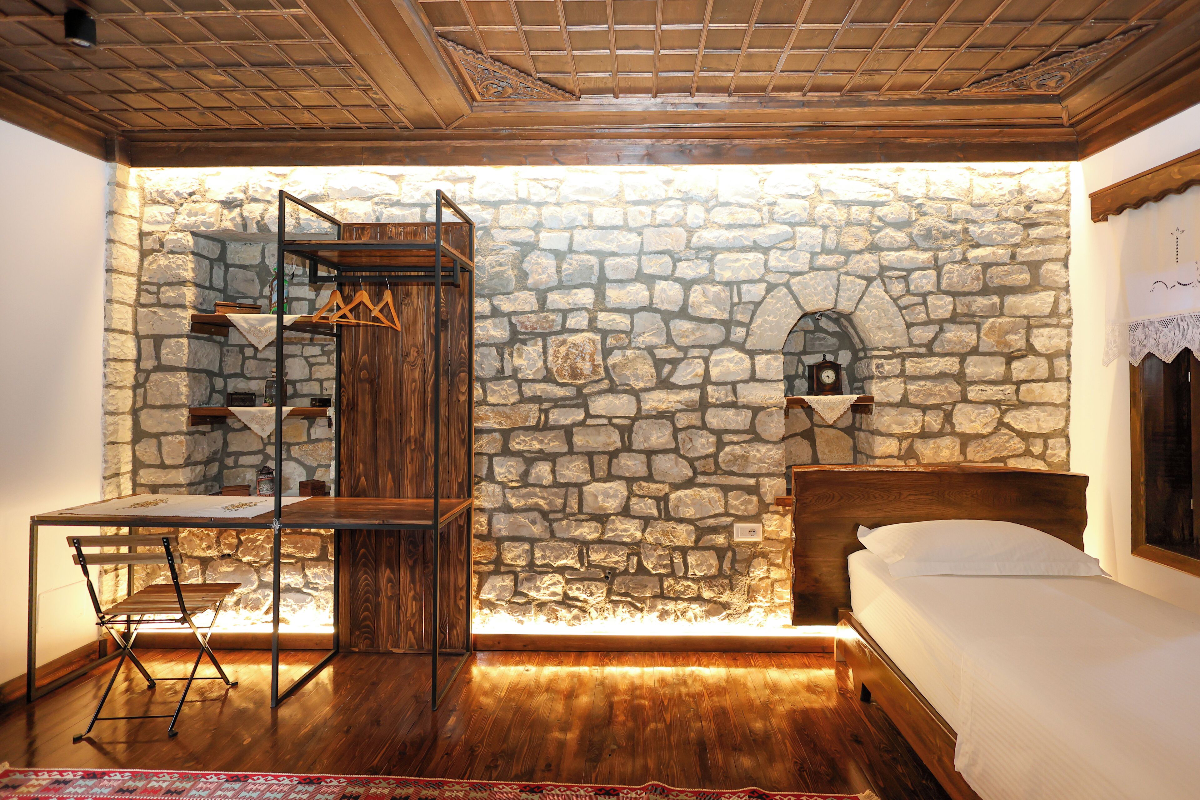 A room with a stone wall, a wooden bed, a desk with a chair, and a small clock on the wall.