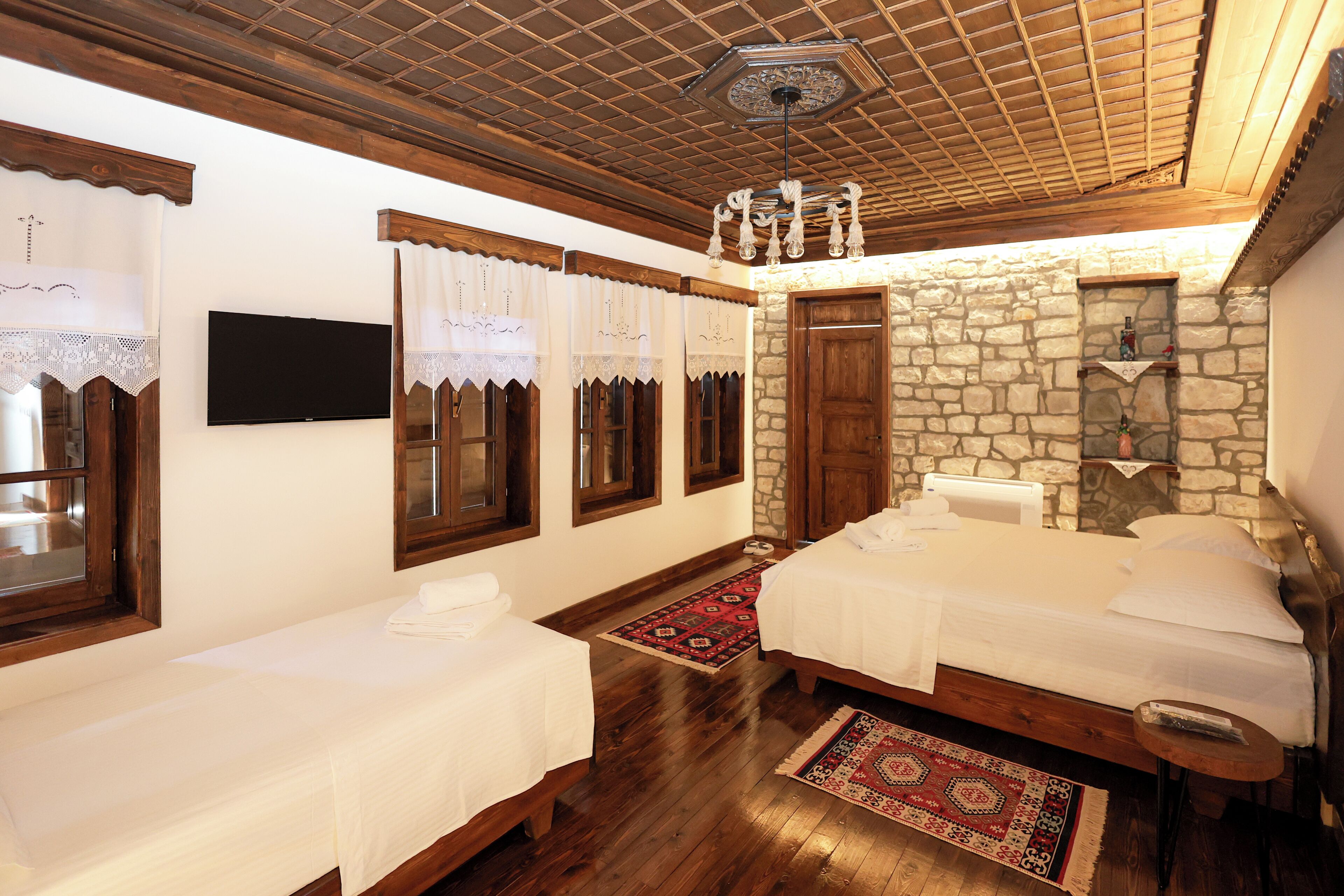 A hotel room with two beds, a stone wall, wooden beams, a TV, and a red and white patterned rug.