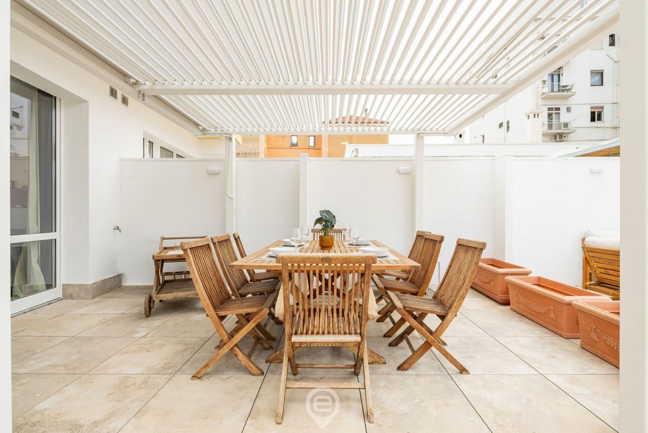 A covered outdoor dining area with a table and chairs, surrounded by white walls and a tiled floor.