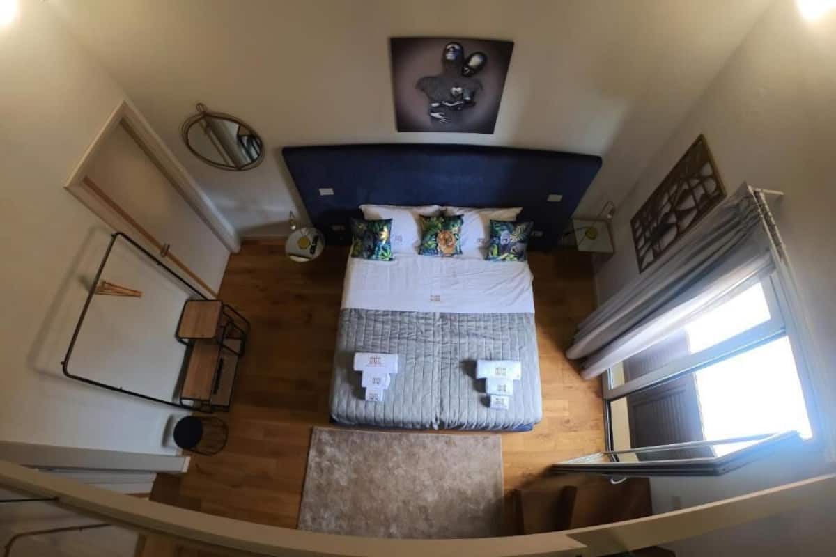 Een slaapkamer met een bed, kussens, een nachtkastje, een spiegel en een raam.