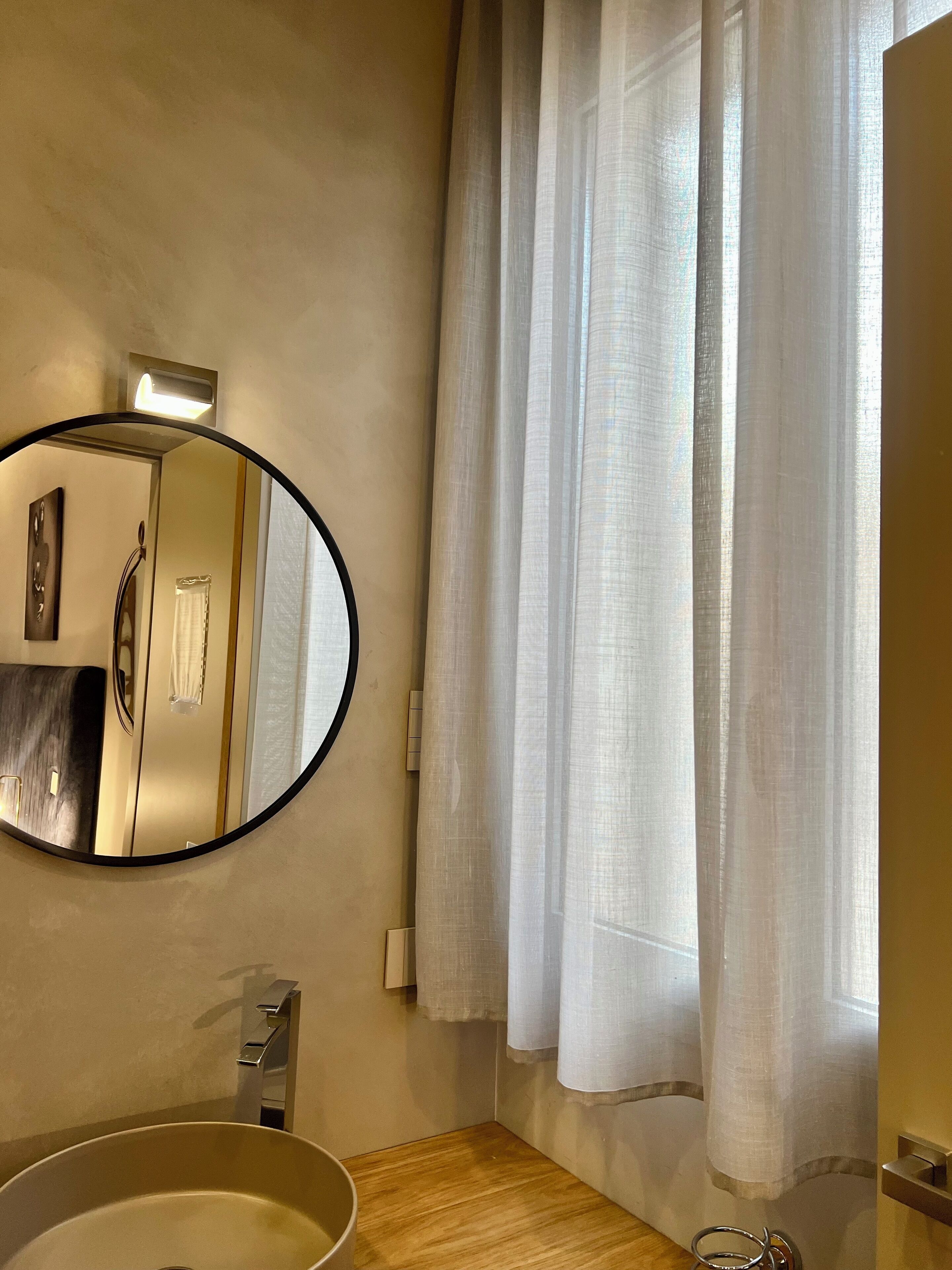 Un baño con un espejo redondo, un lavabo, un perchero para toallas y una ventana con cortinas traslúcidas.