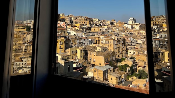 Blick aus einem Fenster auf dicht bebautes Stadtgebiet mit unterschiedlichen Bauweisen und blauem Himmel.