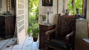 Una habitación con suelo de baldosas, un armario de madera, una silla con cojín estampado, una mesa con lámpara y una ventana con vistas a plantas.