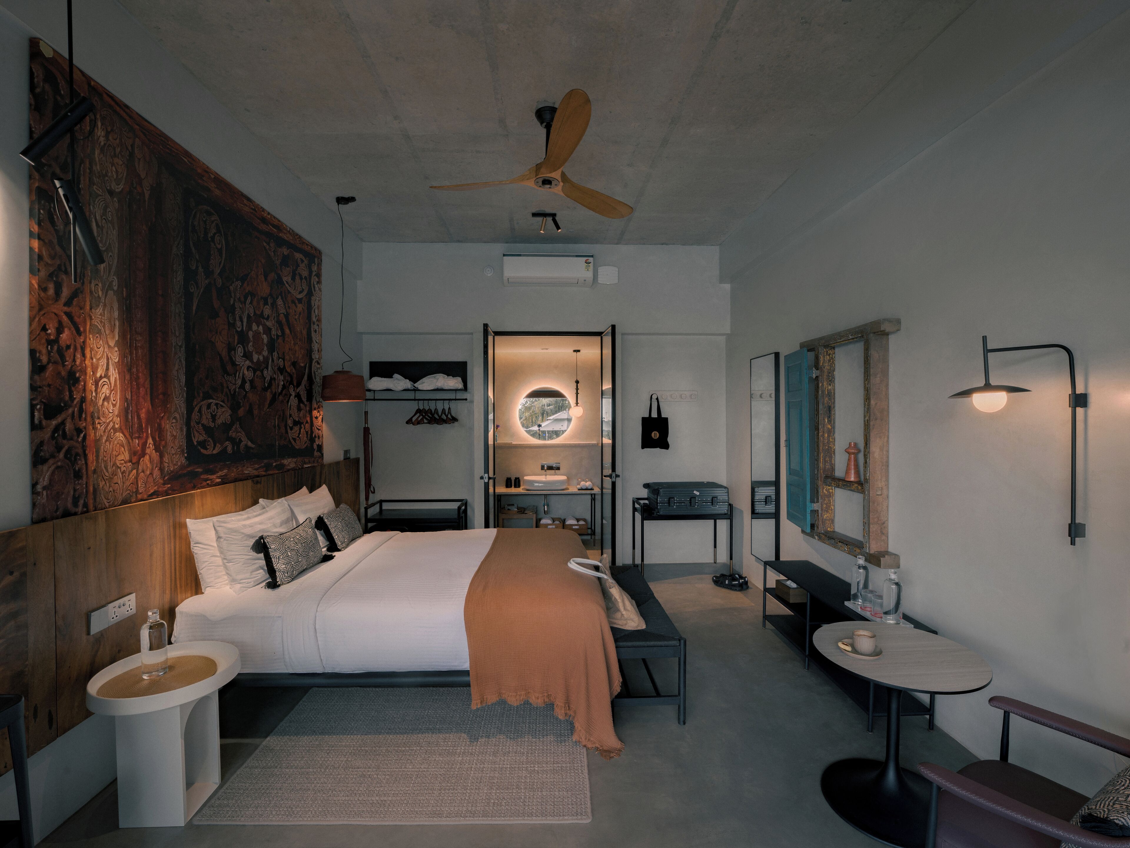 A modern hotel room with a large bed, a desk, a chair, a small table, a mirror, and a ceiling fan.