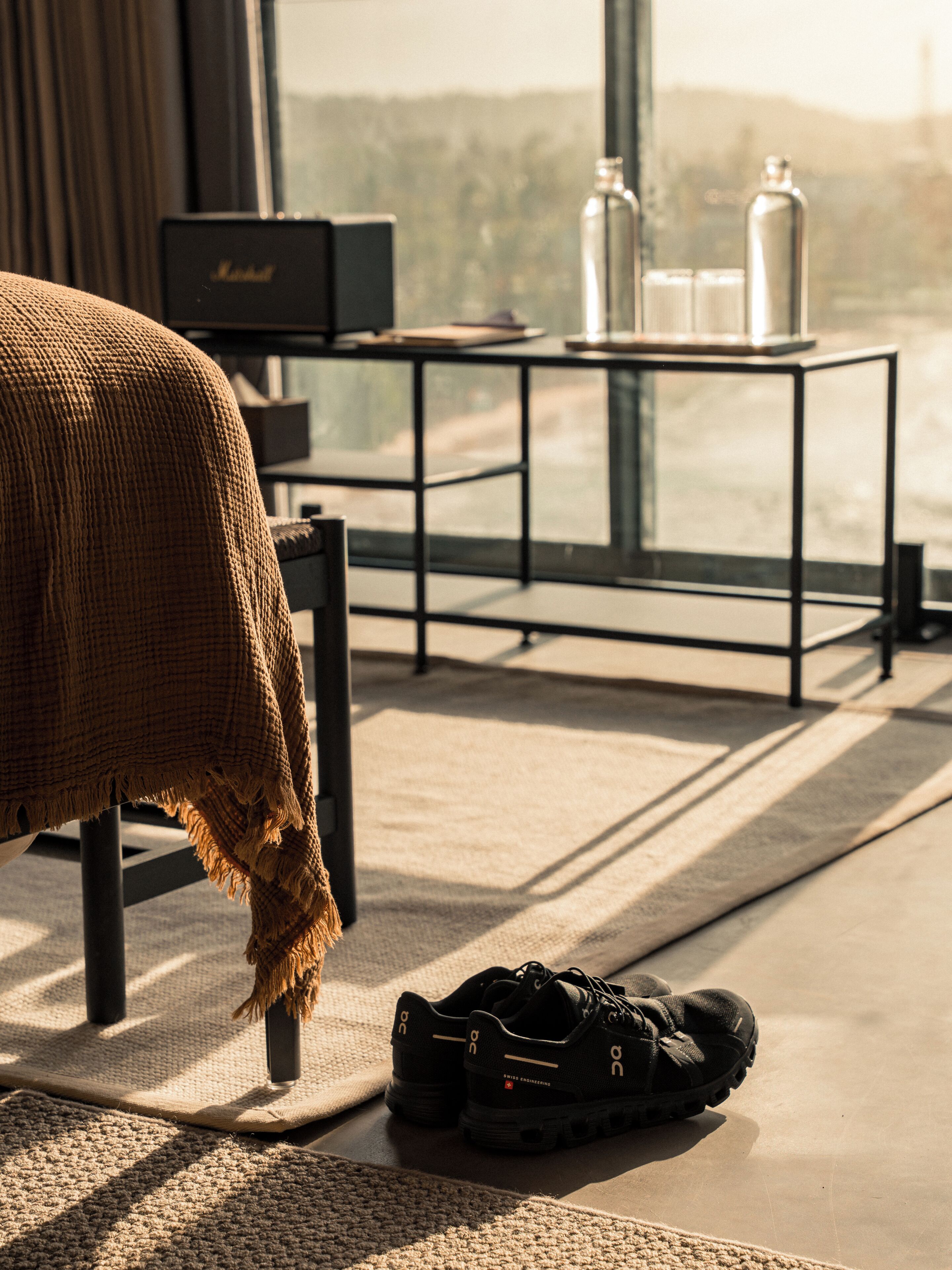 A room with a bed, a pair of shoes on the floor, a small table with bottles, and a view of the outdoors through a window.
