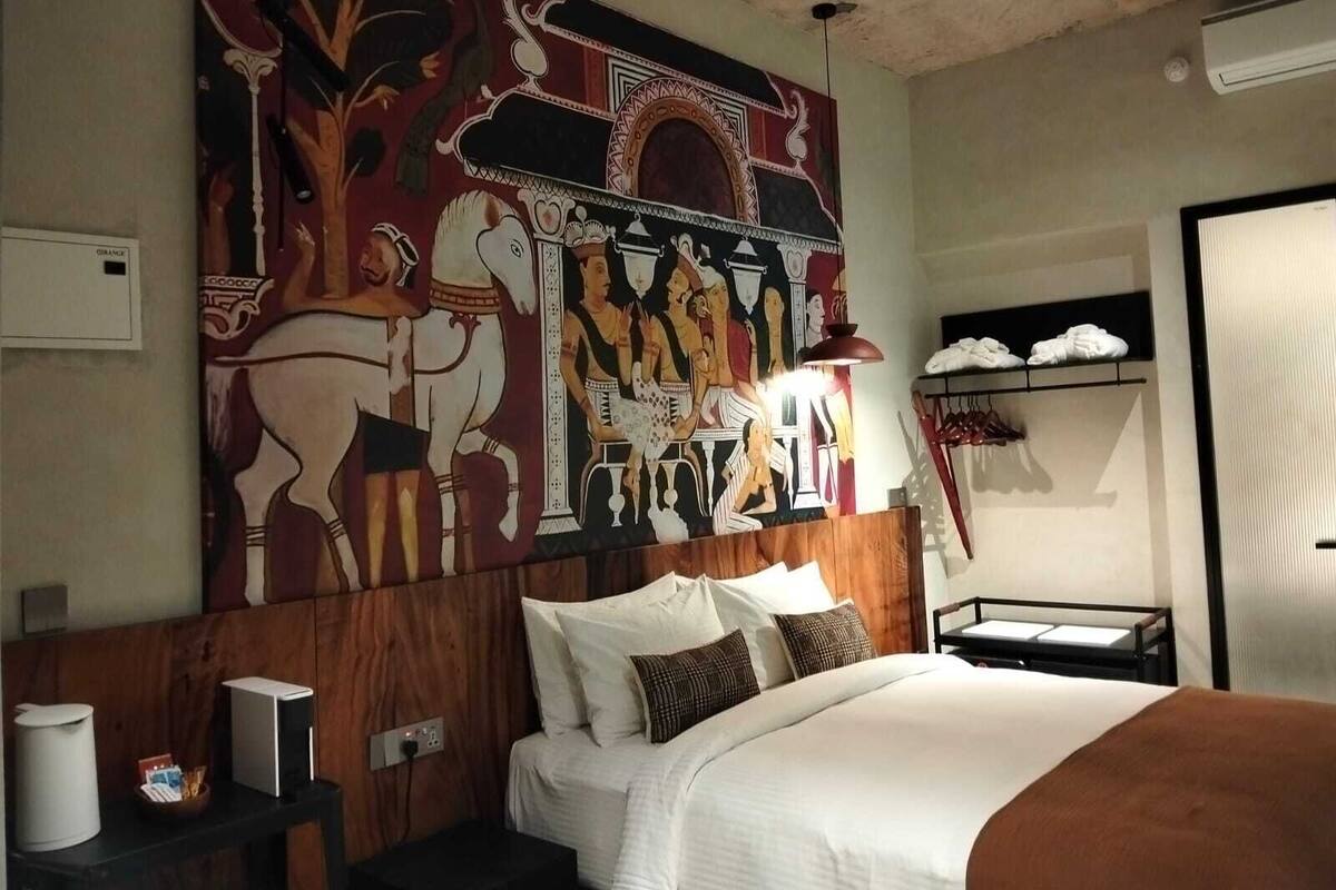 Une chambre d’hôtel avec une grande fresque murale, un lit avec une literie blanche, une tête de lit en bois, une table de chevet avec une lampe et une armoire contenant des vêtements suspendus.