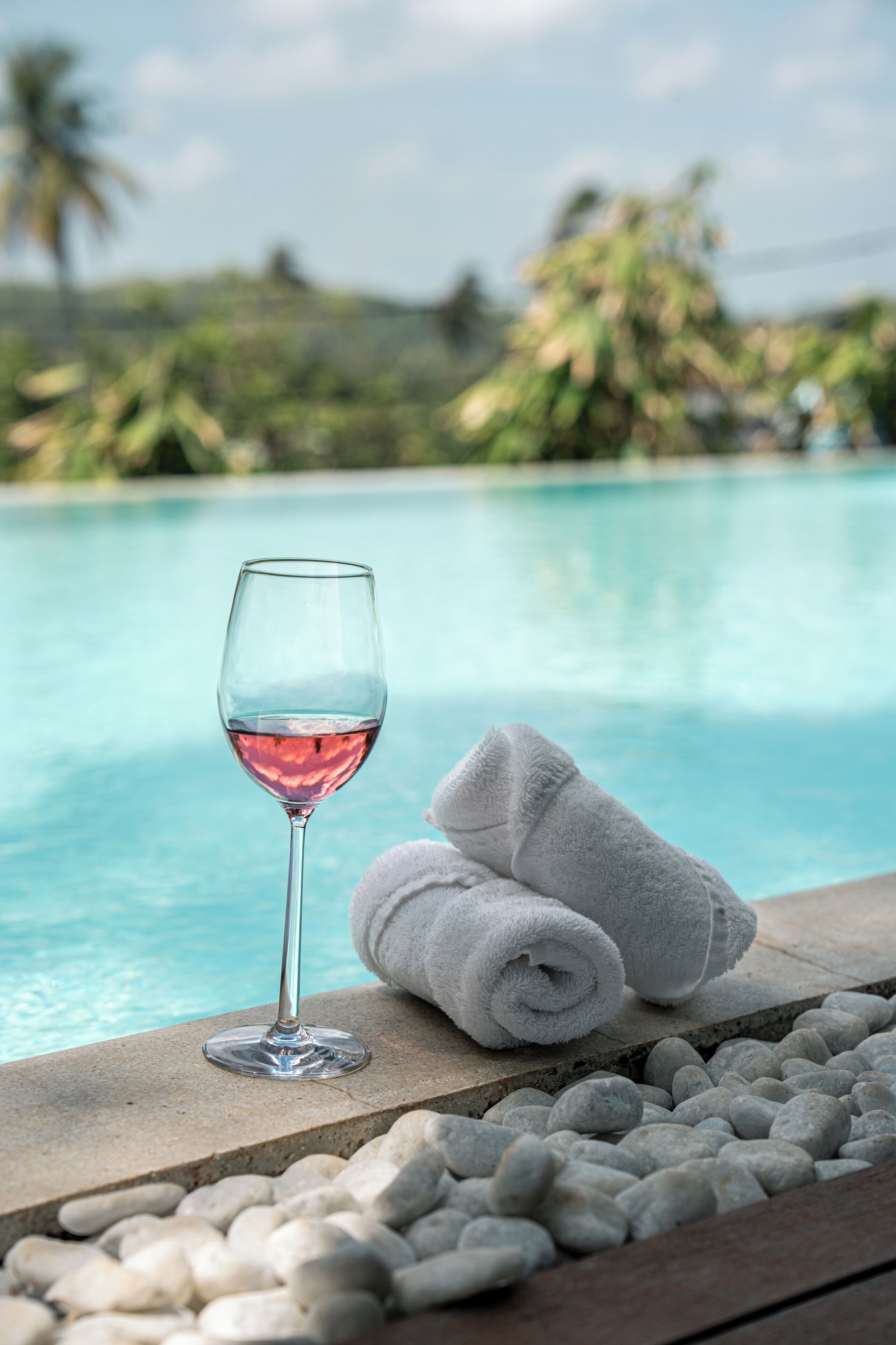 A glass of red wine and two rolled towels by a poolside.