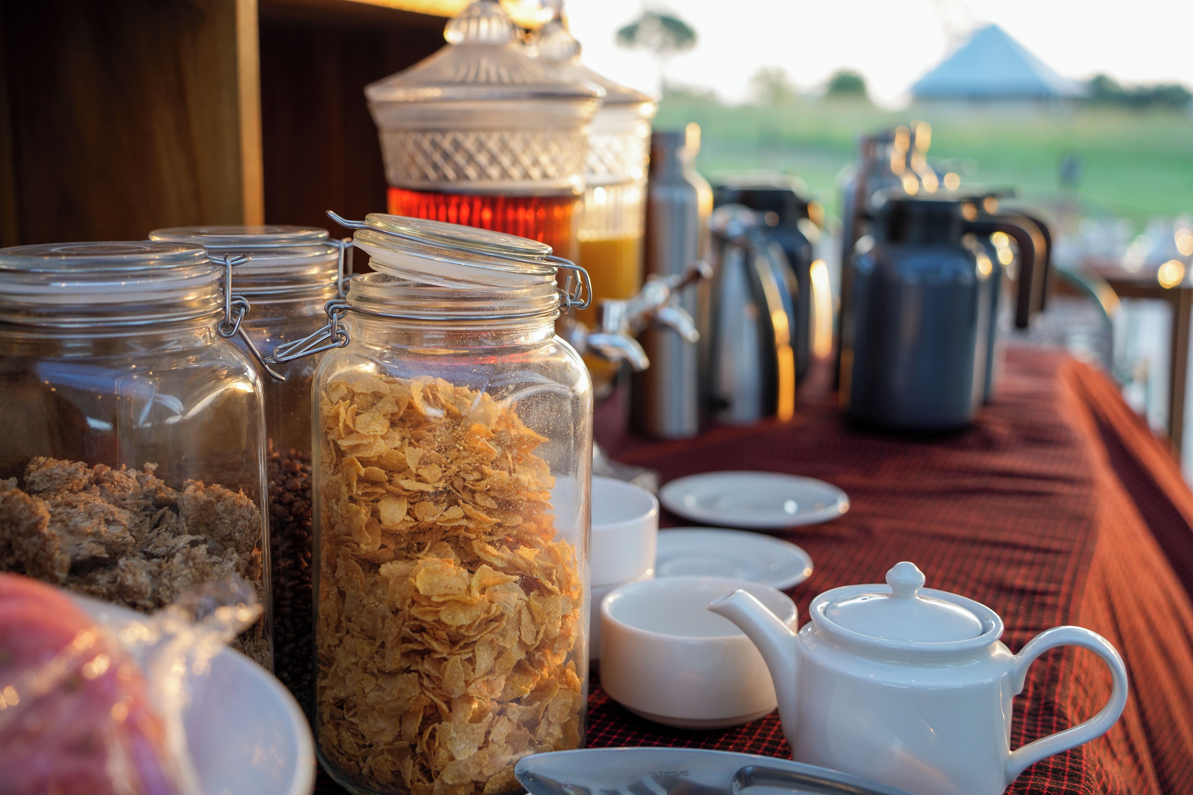 A breakfast setup with jars of granola and cereal, a teapot, cups, and a kettle on a table.