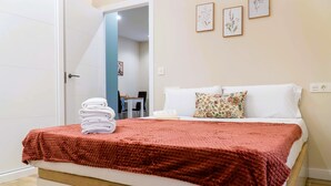 A neatly made bed with a red blanket, white pillows, and a floral throw pillow. There is a stack of white towels on the bed. The room has a white door, a light blue wall, and a small table with chairs in the background. - Apartamentos Doña Berenguela by gaiarooms (León)