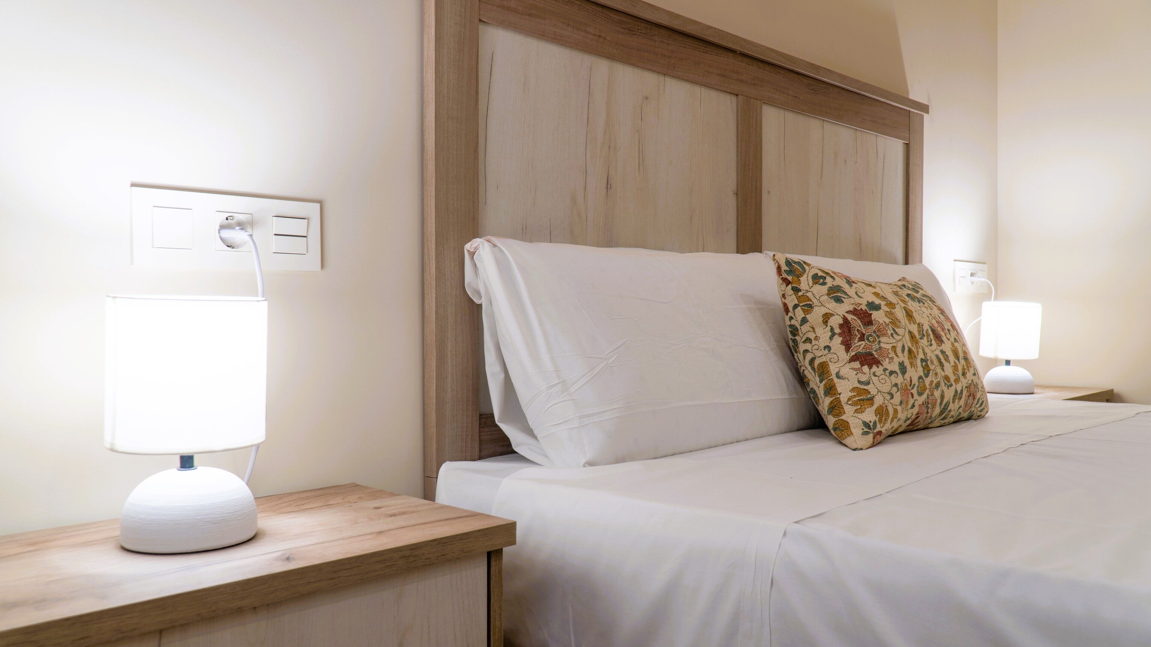 Una cama bien hecha con sábanas blancas y una almohada estampada, flanqueada por dos mesitas de noche con lámparas blancas.