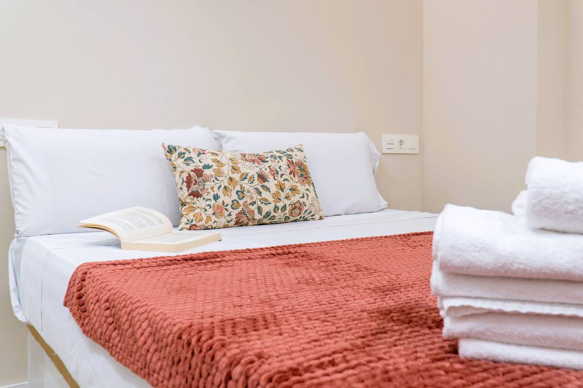 A neatly made bed with white linens, a colorful floral pillow, a red knitted throw, and a stack of white towels.