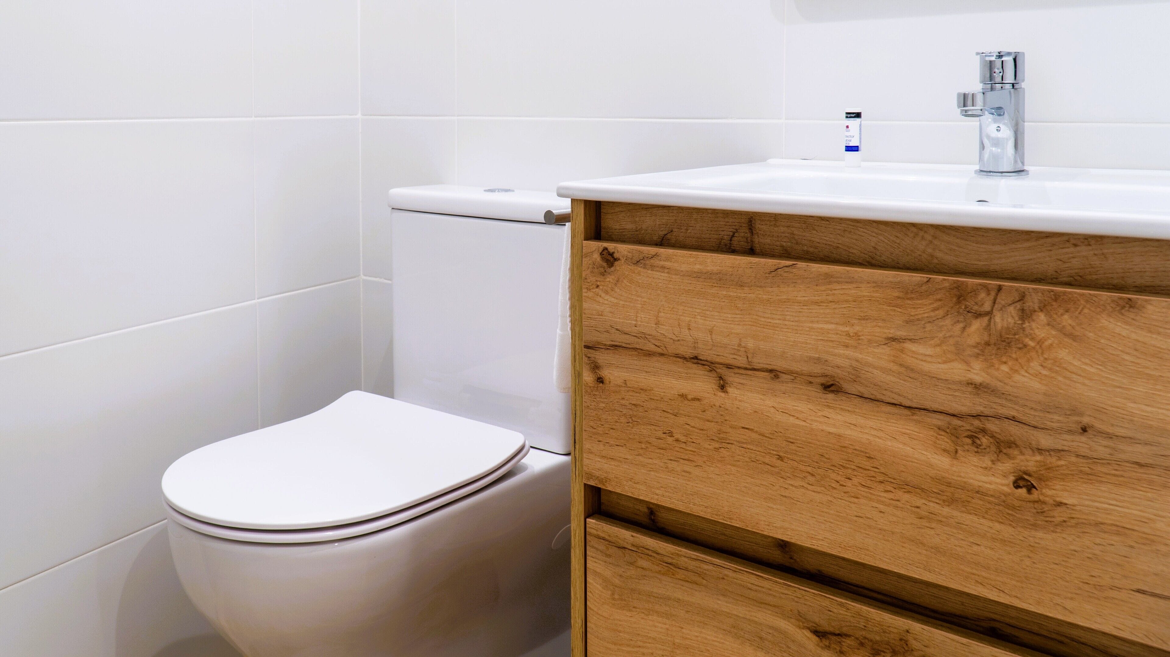 Un baño con inodoro blanco, tocador de madera con lavamanos y pared con azulejos blancos.