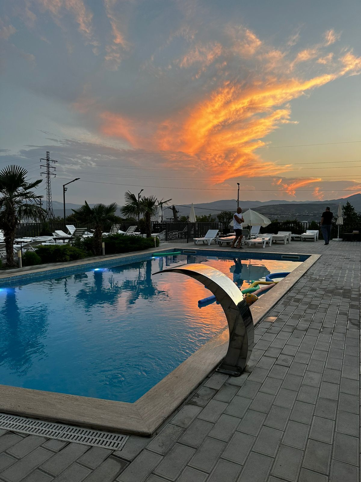 Una piscina con sillas de descanso y sombrillas, rodeada de palmeras y vistas a montañas distantes.